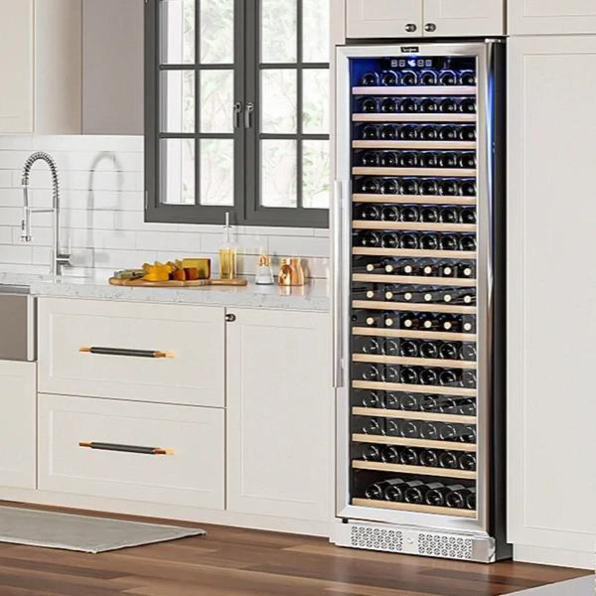 A tall stainless steel wine refrigerator with a glass door stands next to white kitchen cabinets in a modern kitchen. The interior is illuminated with blue light and contains multiple shelves of wine bottles.