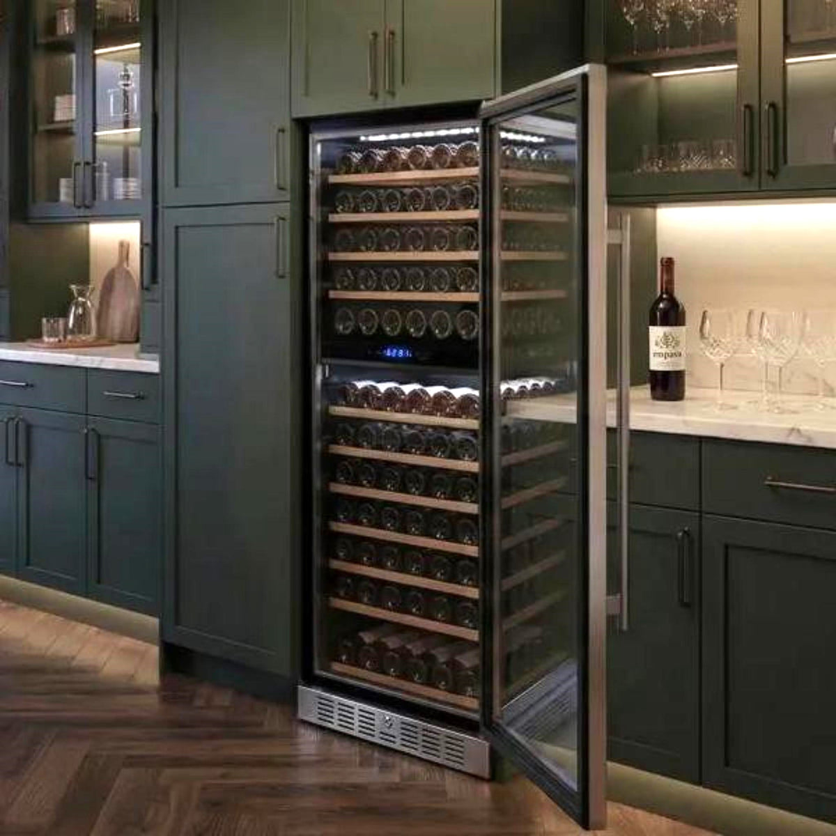 A stainless steel wine refrigerator with its door open, showing multiple shelves of wine bottles inside, set within a dark green kitchen cabinetry environment.