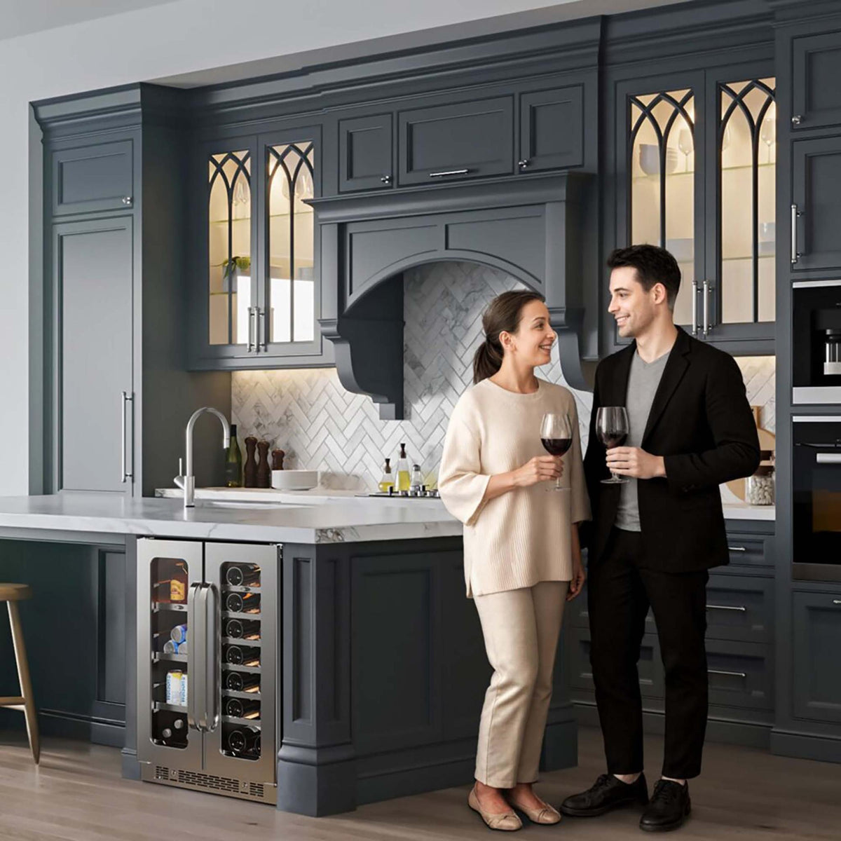 A man and a woman stand in a modern dark gray kitchen, each holding a wine glass. The kitchen features dark gray cabinets, a white marble island, a built-in whynter-BSR-6020FDS-60-can-20-bottle-wine-cooler-refrigerator